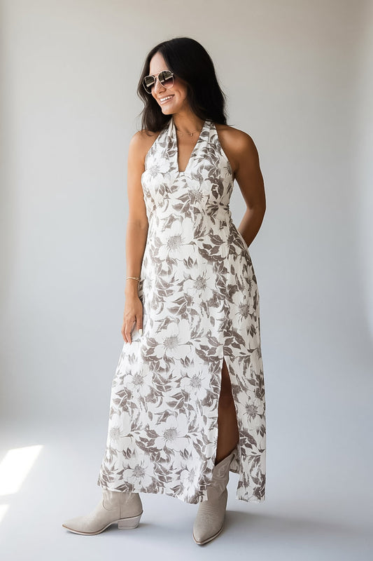 A woman modeling a sleeveless halter-neck maxi dress with a brown and beige floral pattern on a white background, paired with beige ankle boots, set against a plain light gray studio backdrop.