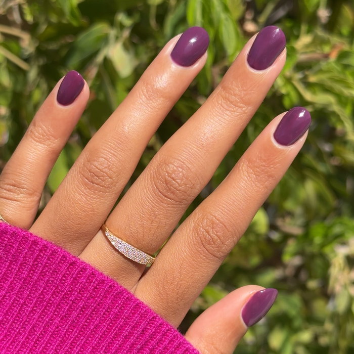 Hand with smooth rounded nails painted in Who Likes Raisins? polish against green foliage background
