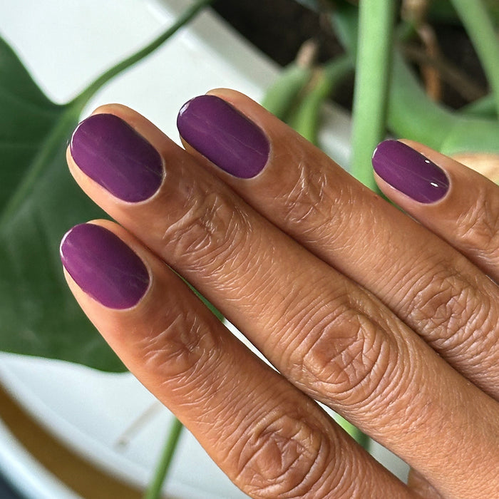 Hand with short rounded nails painted in Olive and June Who Likes Raisins? gel polish against blurred green plant background