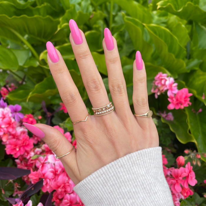 Your Favorite Suit bright peony pink gel polish manicure on hand with gold rings against lush green and pink floral background